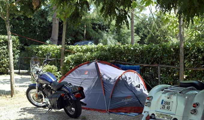Campeggio con tenda, moto e un veicolo a tre ruote parcheggiati all'ombra degli alberi.